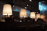 A row of warmly lit table lamps placed on a long wooden table, creating a cozy atmosphere in a dimly lit room. The table also features some decorative elements like small plants and a variety of objects, including a QR code on a stand. There is ambient light in the background and a sparkling decoration on the right side of the image.