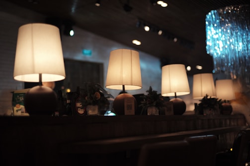 A row of warmly lit table lamps placed on a long wooden table, creating a cozy atmosphere in a dimly lit room. The table also features some decorative elements like small plants and a variety of objects, including a QR code on a stand. There is ambient light in the background and a sparkling decoration on the right side of the image.