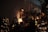 A cozy indoor setting with several illuminated Christmas trees and decorations. The scene is warm with a dimly lit ambience, featuring a large teddy bear perched on a shelf and people gathered at tables, suggesting a festive atmosphere.