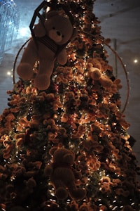 A large Christmas tree is decorated with numerous teddy bears and warm string lights. The bears vary in size and are positioned all over the tree, creating a cozy and whimsical appearance. The setting is illuminated by the glow from the lights, casting a soft and enchanting ambiance.