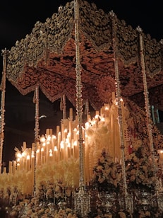 An ornate religious float, richly decorated with intricate patterns and flowers, holds numerous lit candles. The canopy and structural details are lavishly adorned in gold and dark hues, creating a dramatic and solemn atmosphere in the night setting.