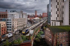 An urban scene featuring a train traveling on elevated tracks through a cityscape filled with a mix of modern and historic buildings. The sky is overcast, adding a dramatic backdrop. Brick structures with aged appearances contrast with sleek, contemporary high-rises, creating a blend of architectural styles.