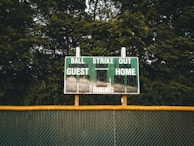 A baseball scoreboard is positioned against a backdrop of dense green trees. The scoreboard displays sections for balls, strikes, outs, guest score, and home score, with the numbers unlit. A chain-link fence with a yellow safety cap runs horizontally beneath the scoreboard.