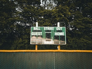 A baseball scoreboard is positioned against a backdrop of dense green trees. The scoreboard displays sections for balls, strikes, outs, guest score, and home score, with the numbers unlit. A chain-link fence with a yellow safety cap runs horizontally beneath the scoreboard.