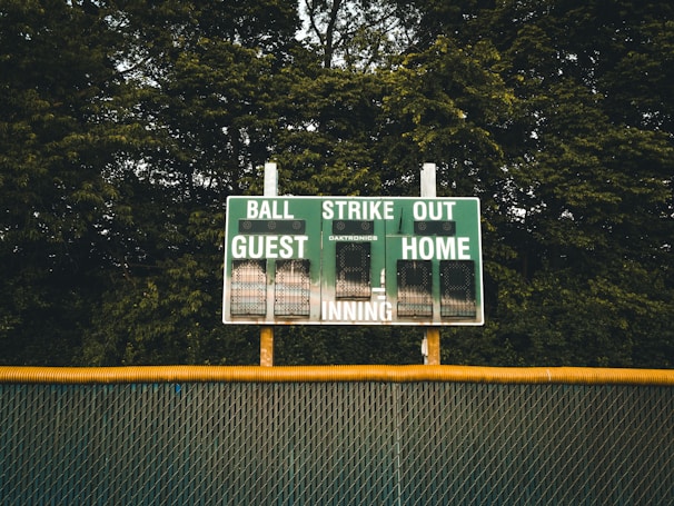 A baseball scoreboard is positioned against a backdrop of dense green trees. The scoreboard displays sections for balls, strikes, outs, guest score, and home score, with the numbers unlit. A chain-link fence with a yellow safety cap runs horizontally beneath the scoreboard.