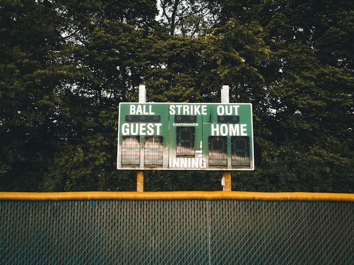 A baseball scoreboard is positioned against a backdrop of dense green trees. The scoreboard displays sections for balls, strikes, outs, guest score, and home score, with the numbers unlit. A chain-link fence with a yellow safety cap runs horizontally beneath the scoreboard.