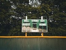A baseball scoreboard is positioned against a backdrop of dense green trees. The scoreboard displays sections for balls, strikes, outs, guest score, and home score, with the numbers unlit. A chain-link fence with a yellow safety cap runs horizontally beneath the scoreboard.