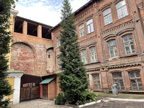 A historic brick building with arched windows and decorative patterns on the facade stands next to a section of exposed brick with arches. A tall evergreen tree is planted in front of the building. The building features a combination of well-preserved and worn elements, indicating age and character. A metal gate and a patch of cobblestones are visible in the foreground.