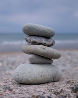 A stack of five smooth, gray stones balanced on top of each other against a blurry seascape backdrop. The stones vary in size, with the largest at the bottom and the smallest at the top. The scene conveys a sense of tranquility and balance.