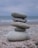A stack of five smooth, gray stones balanced on top of each other against a blurry seascape backdrop. The stones vary in size, with the largest at the bottom and the smallest at the top. The scene conveys a sense of tranquility and balance.