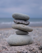 A stack of five smooth, gray stones balanced on top of each other against a blurry seascape backdrop. The stones vary in size, with the largest at the bottom and the smallest at the top. The scene conveys a sense of tranquility and balance.