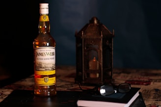 A bottle of James Webb Blended Scotch Whisky is positioned prominently in the foreground, with a decorative metal lantern containing a candle to its right. In front of the bottle lies a closed book with a pair of glasses resting on it. The setting appears to be on a patterned surface, likely a tablecloth, in a dimly lit room with a dark background.