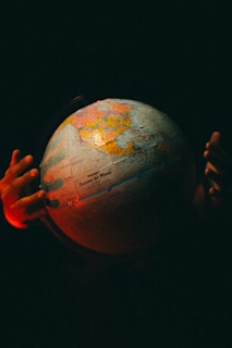 Hands gently hold a globe with highlighted continents in dim lighting. The scene is tinged with a soft, warm glow and is set against a dark background, creating a sense of mystery and focus on the globe itself.