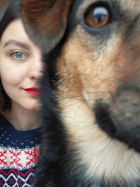 A close-up portrait featuring a woman's face alongside a dog's face. The woman has bright blue eyes and red lipstick, and is wearing a blue sweater with red and white snowflake patterns. The dog's face partially obscures the woman's, with its eye prominently visible and ear upright.