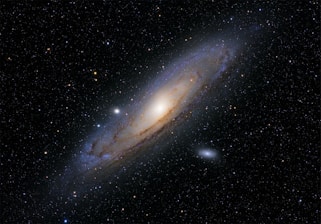 A large spiral galaxy is prominently featured against a backdrop of countless stars. The galaxy has a bright central core with spiraling arms containing dust lanes and clusters of stars. Some smaller, faint celestial objects are visible nearby, enhancing the depth of the scene.