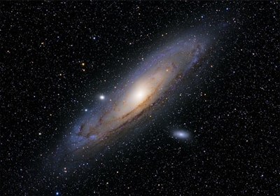 A large spiral galaxy is prominently featured against a backdrop of countless stars. The galaxy has a bright central core with spiraling arms containing dust lanes and clusters of stars. Some smaller, faint celestial objects are visible nearby, enhancing the depth of the scene.