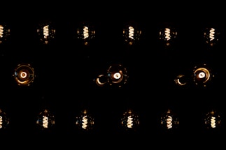 A series of vintage-style Edison light bulbs are arranged in a grid pattern against a dark background. The filaments inside the bulbs emit a warm, orange-yellow glow, highlighting their intricate spirals. The contrast between the dark surrounding and the bright bulbs creates a dramatic visual effect.