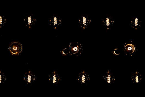 A series of vintage-style Edison light bulbs are arranged in a grid pattern against a dark background. The filaments inside the bulbs emit a warm, orange-yellow glow, highlighting their intricate spirals. The contrast between the dark surrounding and the bright bulbs creates a dramatic visual effect.