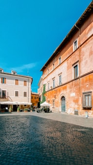 A quaint, sunlit courtyard with cobblestone pavement, surrounded by historic European buildings with warm-toned walls. The scene includes outdoor dining setups with umbrellas and potted plants, and vibrant blue skies above.