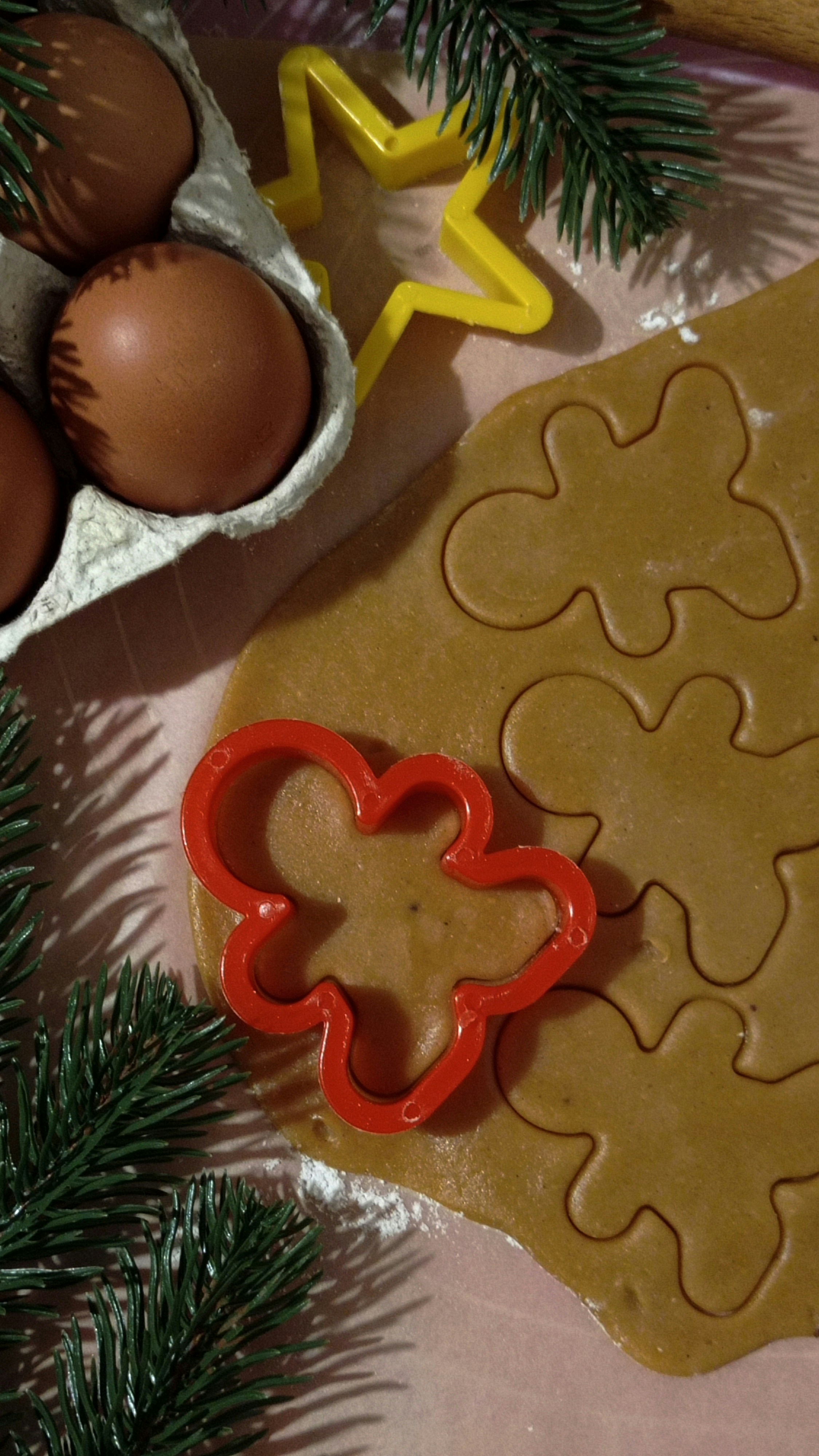 A baking scene featuring rolled-out cookie dough with several gingerbread cutouts. A red butterfly-shaped cookie cutter rests on the dough, near cookie shapes. Next to it, there is a carton containing brown eggs. In the upper section, a yellow star-shaped cutter is visible, and the edges of pine branches encircle the scene, adding a festive touch.