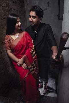 A couple is standing on a narrow staircase. The woman is dressed in a red saree with a patterned blouse and traditional jewelry, while the man is wearing a dark-colored outfit. They are looking at each other with affectionate expressions. The surroundings include worn-out walls that add a rustic charm to the setting.