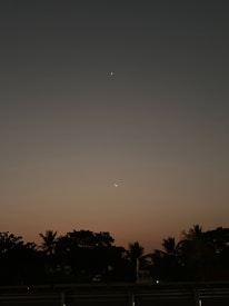 A serene evening sky transitioning into night, with a faint crescent moon and another celestial body visible against a gradient of blue to dusky pink. Silhouetted trees line the horizon, adding a peaceful and tranquil ambiance.