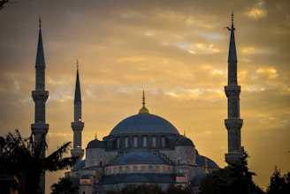 A large mosque with prominent minarets stands silhouetted against a golden sky. The structure features an expansive dome and smaller accompanying semi-domes, indicating traditional Islamic architecture. The sky is filled with clouds, and a bird is flying near one of the minarets.