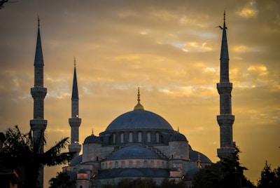 A large mosque with prominent minarets stands silhouetted against a golden sky. The structure features an expansive dome and smaller accompanying semi-domes, indicating traditional Islamic architecture. The sky is filled with clouds, and a bird is flying near one of the minarets.