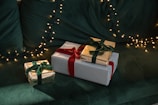 Three wrapped gifts are arranged on a green, plush surface. The gifts have gold and striped wrapping paper, and are tied with green and red velvet ribbons. In the background, there are warm white fairy lights adding a festive ambiance.