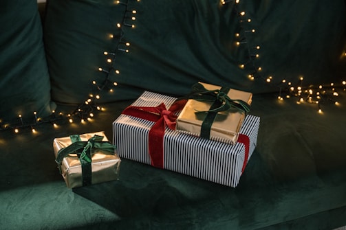 Three wrapped gifts are arranged on a green, plush surface. The gifts have gold and striped wrapping paper, and are tied with green and red velvet ribbons. In the background, there are warm white fairy lights adding a festive ambiance.