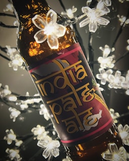 A bottle of beer labeled 'India Pale Ale' is surrounded by decorative lights shaped like white flowers. The background is dark, highlighting the warm amber color of the bottle and the glowing lights.