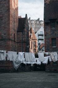 A row of laundry hung to dry between two historic buildings, with a backdrop of more buildings in the distance. The clothes are predominantly white and include various garments such as shirts and towels. The architecture features red brick and stone with pointed roofs and chimneys.