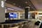 An airport baggage claim area with a conveyor belt and luggage. Digital screens displaying information are mounted above the conveyor belt. The space is illuminated with overhead lighting, and signage directs passengers to taxis and exits.