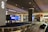 An airport baggage claim area with a conveyor belt and luggage. Digital screens displaying information are mounted above the conveyor belt. The space is illuminated with overhead lighting, and signage directs passengers to taxis and exits.