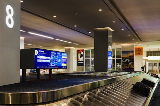 An airport baggage claim area with a conveyor belt and luggage. Digital screens displaying information are mounted above the conveyor belt. The space is illuminated with overhead lighting, and signage directs passengers to taxis and exits.