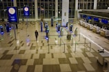 An airport terminal with a mostly empty floor. There are self-check-in kiosks with glowing blue lights, surrounded by metal barriers. A single person walks through the space, and a few individuals are seated near the large glass windows. Advertisements are displayed on digital screens mounted on tall poles, and airline check-in counters are visible in the background.