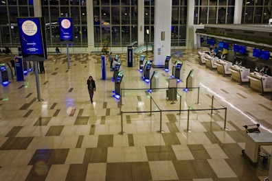 An airport terminal with a mostly empty floor. There are self-check-in kiosks with glowing blue lights, surrounded by metal barriers. A single person walks through the space, and a few individuals are seated near the large glass windows. Advertisements are displayed on digital screens mounted on tall poles, and airline check-in counters are visible in the background.