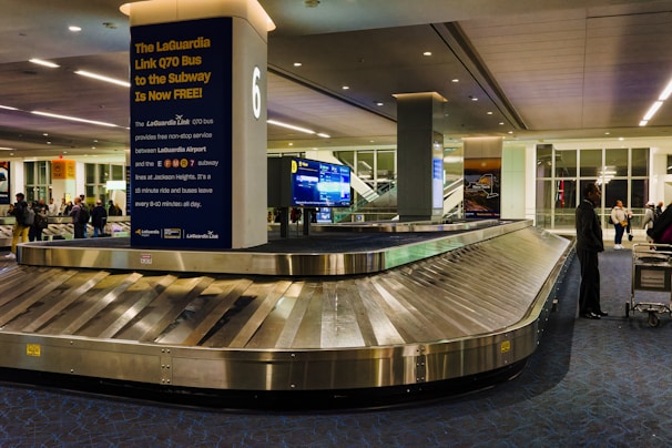 A quiet baggage claim area in an airport with modern lighting and architecture. A large carousel with the number 6 displayed is positioned in the center. There is a sign promoting the free LaGuardia Link Q70 bus service to the subway. Several people are around the area, some with luggage on carts.