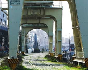 A large industrial structure spans across a cobblestone pathway with unused railway tracks. The metal beams, painted in beige, have various plaques attached. In the background, a cityscape with historic buildings is visible, partially obscured by mist.