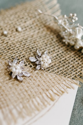 A pair of intricate pearl and crystal earrings are placed on a textured burlap fabric, displaying a delicate and elegant style. The earrings have a floral design, with small pearls surrounded by sparkling crystals. In the background, there are subtle hints of other decorative items, adding to the sophisticated arrangement.