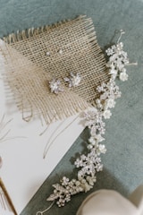 A decorative arrangement with a delicate floral hair accessory adorned with pearls and intricate crystal details placed on a textured burlap surface. The backdrop includes a soft green fabric and a piece of white paper.