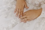 Hands with neatly manicured nails are placed on a white floral-patterned fabric, possibly a dress, and a gold ring is visible on one finger.