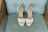 A pair of elegant white high-heeled shoes with ankle straps is placed on a light blue cushioned chair with wooden wicker arms. The shoes feature a crossover design on the toe area.