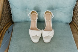 A pair of elegant white high-heeled shoes with ankle straps is placed on a light blue cushioned chair with wooden wicker arms. The shoes feature a crossover design on the toe area.