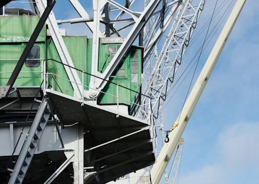 A close-up of a large industrial crane with a green cabin and white metallic structure. The crane features a complex network of beams and cables against a clear blue sky.