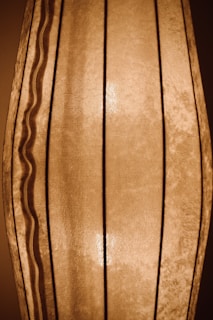 A close-up view of a vertically pleated fabric lampshade with a soft, sepia-toned light emanating from behind it. There are both straight and wavy lines forming the structure, giving it a distinctive texture and pattern.