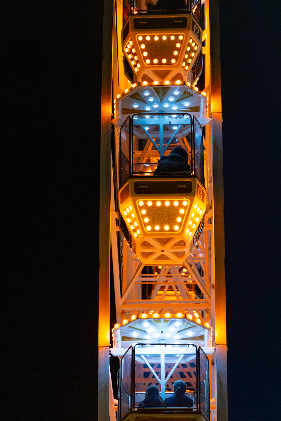 The image depicts a large Ferris wheel illuminated by orange and white lights against a dark night sky. The Ferris wheel is composed of multiple levels, each adorned with colorful lights that create a vibrant spectacle. Atop the structure, there are two individuals seated in seats facing forward, likely enjoying the view from their elevated vantage point.