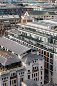 Aerial view of a densely built urban area featuring modern high-rise and mid-rise buildings with large glass windows and flat roofs. The buildings are closely packed together, showcasing a blend of architectural styles. Rooftop terraces with greenery are visible, suggesting environmental considerations in the architectural design.