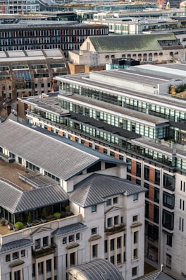 Aerial view of a densely built urban area featuring modern high-rise and mid-rise buildings with large glass windows and flat roofs. The buildings are closely packed together, showcasing a blend of architectural styles. Rooftop terraces with greenery are visible, suggesting environmental considerations in the architectural design.