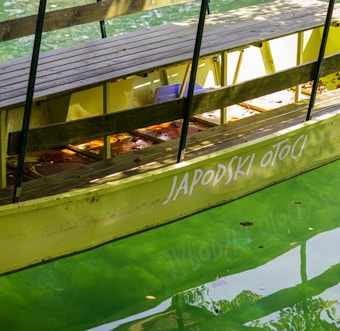 A yellow-green wooden boat with benches is docked on a bright green body of water. The boat has the words 'JAPODSKI OTOCI' painted on its side in white letters. Sunlight casts shadows and reflections on the water and various surfaces of the boat.